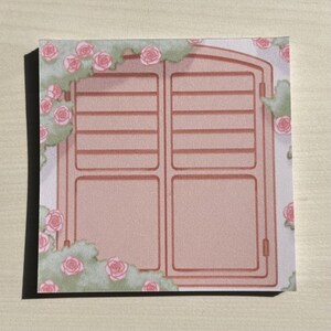 Puede incluir: Una nota adhesiva cuadrada con una ilustración impresa de una ventana de color marrón claro con contraventanas, rodeada de racimos de rosas de color rosa suave y follaje verde apagado. El diseño cubre toda la superficie del papel.