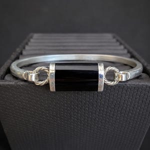 May include: A silver bangle bracelet featuring a rectangular black stone centerpiece. The stone is flanked by two twisted silver rings that connect the central piece to the smooth, curved silver band, creating a polished and structured design.