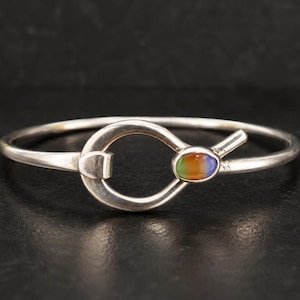 May include: A silver bangle bracelet featuring a central loop design that functions as a clasp. The clasp is accented with a small, oval-shaped cabochon stone displaying a gradient of colors including orange, green, and blue, set in a smooth metal frame.