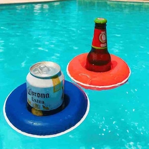 May include: A blue inflatable drink holder containing a silver Corona Extra aluminium can and a red inflatable drink holder containing a brown glass beer bottle with a red and white label, both floating on the surface of bright blue pool water.