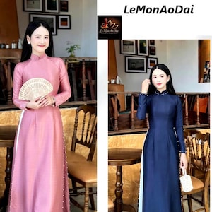 May include: Two women in traditional Vietnamese Áo Dài dresses. One is a soft pink with white trim and pearl accents, the other a deep navy blue with similar details. The dresses have long sleeves and high collars, reflecting Vietnamese culture.