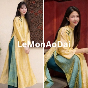 Könnte beinhalten: Ein traditionelles vietnamesisches Ao Dai-Kleid in einem sanften Gelbton, mit hohem Kragen und langen Ärmeln. Das Kleid hat einen geteilten Vorderteil, der eine teal-farbene Hose darunter freigibt. Der Text "LeMonAoDai" ist sichtbar.