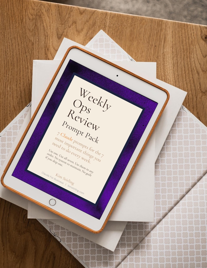 May include: A digital tablet with a brown frame displays the text "Weekly Ops Review Prompt Pack." The screen has a purple border and white background. The tablet rests on a stack of white and patterned paper, set on a wooden surface.