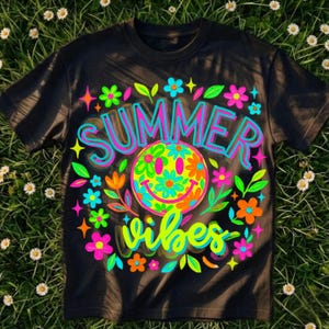 Könnte beinhalten: Schwarzes T-Shirt mit einem farbenfrohen Grafikdesign. Das Design zeigt die Worte "SUMMER VIBES" in einer Neon-Schrift, ein Smiley-Gesicht mit Blumendetails und verschiedene Blumen und Sterne. Das T-Shirt befindet sich auf einem grasbewachsenen Hintergrund.