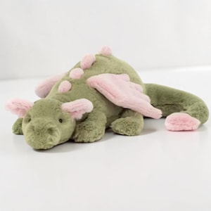 Könnte beinhalten: Ein grünes Plüschdrachenspielzeug mit rosa Akzenten. Der Drache hat rosa Flügel, Ohrendetails und Rückenstacheln. Das Stofftier liegt auf einer weißen Oberfläche und zeigt sein detailliertes Design und sein kuscheliges Aussehen.