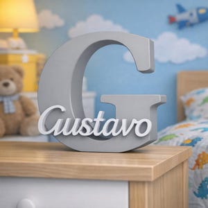 Puede incluir: Una letra "G" gris en 3D con el nombre "Gustavo" en escritura blanca, exhibida sobre una mesita de noche de madera. El entorno del dormitorio incluye un oso de peluche, una lámpara y una cama con un edredón con temática de dinosaurios. El fondo presenta una pared azul con nubes y un avión de juguete.