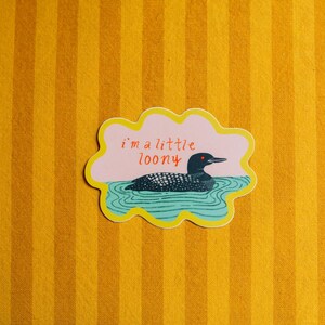 May include: A die-cut sticker with a scalloped yellow border featuring a black and white loon swimming in teal water. The background is light pink with the red text "i'm a little loony" written above the bird. The sticker has a matte finish.