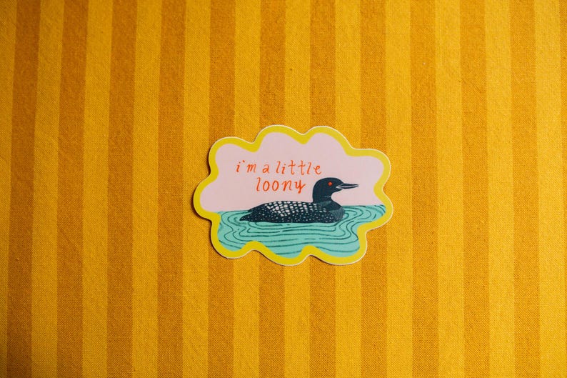 May include: A die-cut sticker with a scalloped yellow border featuring a black and white loon swimming in teal water. The background is light pink with the red text "i'm a little loony" written above the bird. The sticker has a matte finish.
