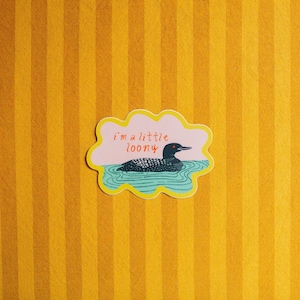 May include: A die-cut sticker with a scalloped yellow border featuring a black and white loon swimming in blue water against a light pink background. The text "i'm a little loony" is printed in red above the bird, which has a bright orange eye.