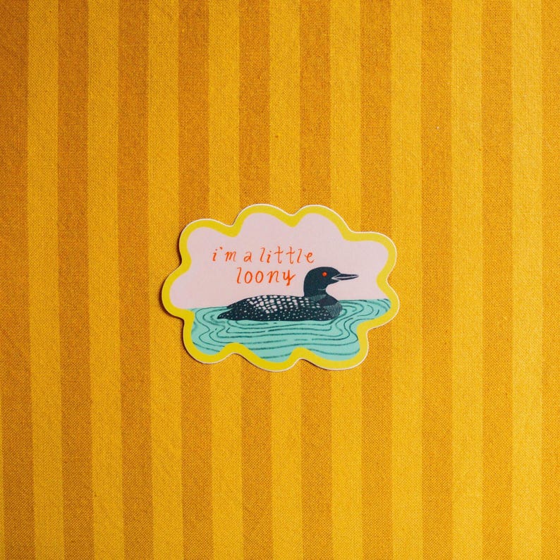 May include: A die-cut sticker with a scalloped yellow border featuring a black and white loon swimming in blue water against a light pink background. The text "i'm a little loony" is printed in red above the bird, which has a bright orange eye.