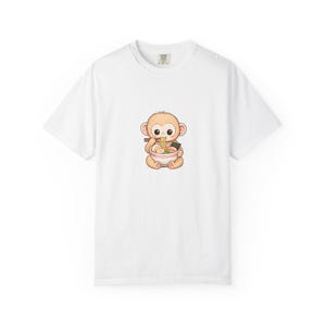 以下が含まれることがあります： ラーメンを食べている漫画の猿が描かれた白いTシャツ。猿は明るい茶色で大きな目を持ち、箸を持っています。ラーメンの丼は白で、赤と緑のデザインです。シャツはクラシックなクルーネックです。