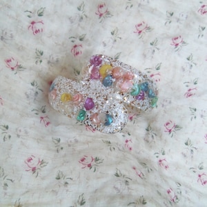 May include: Two white, curved bracelets are adorned with colorful beads and sequins. The bracelets are decorated with a variety of pastel-colored beads, including pink, yellow, blue, and green. The background is a floral patterned fabric.
