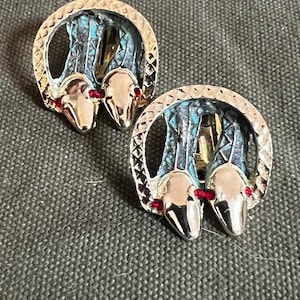 May include: A pair of silver-toned clip-on earrings. Each earring features a circular design with a textured border and a snake head motif. The snakes have red gemstone eyes and are set against a blue background.