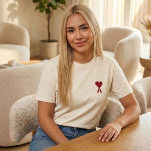 Könnte beinhalten: Cremefarbenes T-Shirt mit einem kleinen roten Herz und dem Buchstaben "A". Die Person, die das T-Shirt trägt, hat blonde Haare und sitzt an einem Holztisch. Der Hintergrund zeigt ein Wohnzimmer mit Möbeln in neutralen Farbtönen.