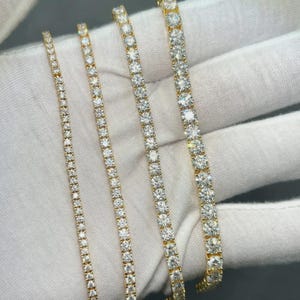May include: Four gold tennis bracelets, each set with clear, sparkling gemstones. The bracelets vary in width, with the gemstones closely set. The bracelets are displayed on a white surface, highlighting their brilliance.