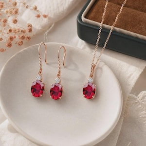 Può includere: Un set di gioielli composto da un paio di orecchini pendenti e una collana con ciondolo abbinata. Ogni pezzo presenta una gemma rosso scuro taglio ovale incastonata in oro rosa, con una piccola pietra chiara rotonda sopra la gemma principale.
