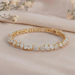 Peut inclure: Un bracelet doré orné de pierres précieuses claires de différentes formes, dont des poires, des émeraudes et des rondes. Le bracelet est présenté sur un fond neutre et doux, mettant en valeur l'élégance du bijou.