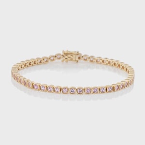 May include: A gold-colored bracelet with a series of square-cut, light pink gemstones. The bracelet has a clasp closure and is displayed on a white background. The gemstones are evenly spaced along the bracelet's length.