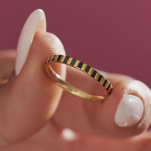 May include: A gold ring with a repeating pattern of black and gold stripes. The ring is held between fingers with white nail polish against a pink background. The ring is a band style.
