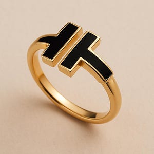 May include: A gold-colored ring with a black enamel inlay in a bold T-shaped design. The ring has a smooth, polished finish and a minimalist aesthetic, suitable for everyday wear.