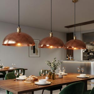 May include: Three copper pendant lights with a hammered texture hang above a wooden dining table. The lights have a dome shape and are suspended by black cords. The dining table is set with plates, glasses, and a loaf of bread, with green velvet chairs.