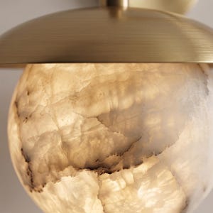 May include: A wall-mounted light fixture featuring a spherical, translucent white stone shade with natural veining patterns. The light is topped with a brushed brass metal dome cap that extends over the top of the illuminated stone sphere.