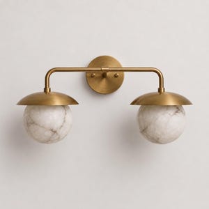 May include: A wall-mounted light fixture featuring a horizontal brass bar attached to a circular backplate. Two brass dome-shaped shades extend from the bar, each housing a spherical white marble glass globe with grey veining patterns.