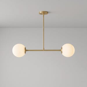 May include: A modern, two-light pendant light fixture. It features a brass finish with two spherical, frosted glass globes. The fixture is suspended from the ceiling by a vertical rod and a horizontal bar.