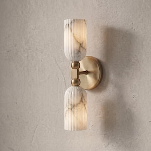 Può includere: Una lampada da parete con due paralumi cilindrici scanalati sovrapposti verticalmente, realizzati in marmo bianco con venature grigie. I paralumi sono collegati da un'asta centrale color ottone e fissati a una piastra circolare a parete.