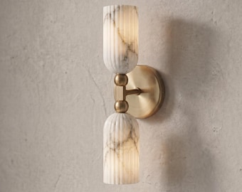 May include: A wall sconce featuring two vertically stacked, fluted cylindrical shades made of white marble with grey veining. The shades are connected by a central brass-toned metal rod and mounted to the wall with a circular brass-toned plate.