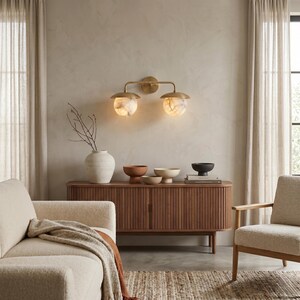 May include: A wall-mounted light fixture featuring two brass arms and two spherical, marbled glass shades that emit a warm glow. The fixture is mounted on a circular brass base and is positioned above a wooden sideboard with vertical slats.