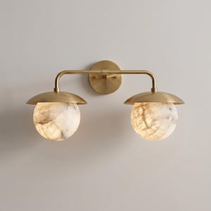 May include: A wall-mounted light fixture featuring a horizontal brass arm and a circular mounting plate. Two downward-facing brass shades hold spherical, translucent white stone globes with grey veining that emit a soft, warm glow.