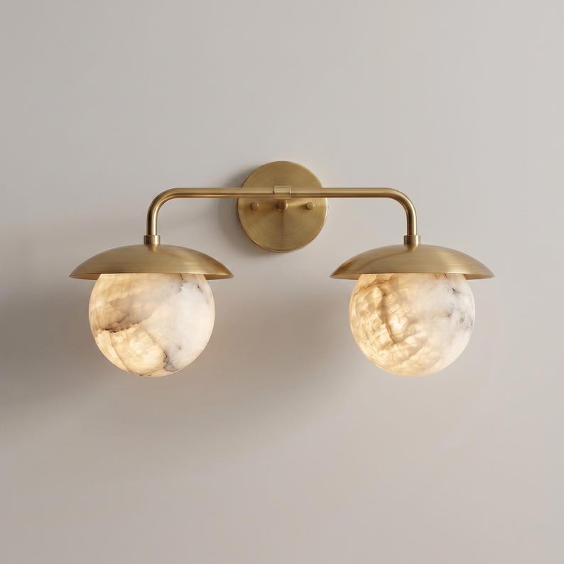 May include: A wall-mounted light fixture featuring a horizontal brass arm and a circular mounting plate. Two downward-facing brass shades hold spherical, translucent white stone globes with grey veining that emit a soft, warm glow.