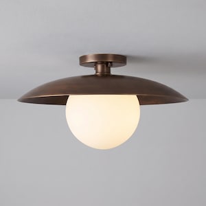 May include: A semi-flush mount ceiling light fixture featuring a wide, dark bronze metal disc shade and a central, spherical white glass globe. The fixture is mounted to a circular bronze base that attaches directly to the ceiling surface.