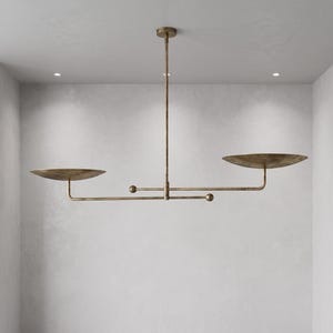 May include: A linear pendant light fixture in an antique brass finish, featuring a central vertical rod connected to a horizontal bar. Two shallow, bowl-shaped discs are positioned at opposite ends, balanced by small spherical weights on the arms.
