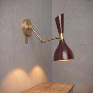 May include: A wall-mounted, adjustable sconce with a burgundy shade and brass accents. The sconce has a circular base and an articulated arm. The shade is a deep burgundy colour, and the fixture is mounted on a grey wall.
