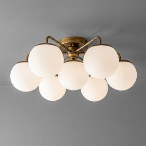 May include: A ceiling light fixture with a brass finish and seven spherical, frosted glass shades. The fixture is mounted on a gray ceiling. The design is modern and provides ambient lighting.