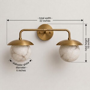 May include: A brass wall light fixture with two white alabaster globes. The fixture has a total width of 22 inches, a total height of 10 inches, and each alabaster globe has a diameter of 6 inches. The design features a central circular mount.