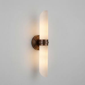 May include: A wall sconce featuring a cylindrical, vertically ribbed frosted glass shade divided into two sections by a central bronze-toned metal band. The fixture is mounted to the wall with a circular bronze-toned base, emitting a warm glow.