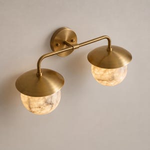 May include: A wall-mounted light fixture featuring a circular brass backplate and two curved arms extending to support two downward-facing lamps. Each lamp has a brushed brass dome shade and a spherical, translucent white stone globe diffuser.