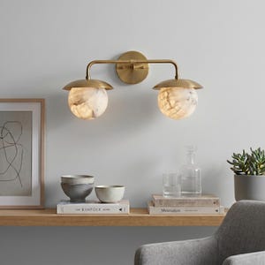 May include: A wall-mounted light fixture featuring a horizontal brass arm attached to a circular backplate. Two downward-facing brass shades hold spherical, translucent white stone globes with natural grey veining, providing illumination.
