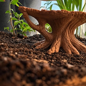 May include: A brown, textured, tree-stump-shaped platform with a flat top and a hollow opening in the centre, resting on a dark soil surface. The object features realistic bark-like detailing and root structures extending from the base.