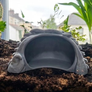 May include: A grey, semi-circular, hollow structure with a textured, rock-like exterior surface and a smooth, arched opening. The object features a layered, rugged design resembling a natural stone formation or a small cave entrance.