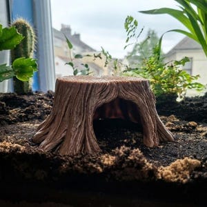 May include: A brown, hollow tree stump ornament with a textured, bark-like surface and a wide, arched opening at the front. The object is positioned on dark soil, featuring deep vertical grooves and ridges that mimic the appearance of natural wood.