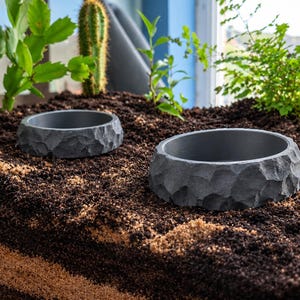 May include: Two circular, dark grey bowls with a textured, chiseled exterior surface that mimics the appearance of natural stone. The bowls are set into dark brown soil, with green plants visible in the background behind the containers.