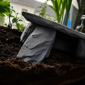 May include: A grey stone-like shelter structure featuring a flat rectangular roof slab supported by three vertical rock-shaped pillars. The object has a textured, layered appearance resembling natural slate and creates a hollow opening underneath.