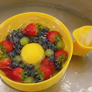 Op de afbeelding: Een felgele ronde fruitwasbak met een verhoogde koepel in het midden en een tuit aan de zijkant. De kom is gevuld met water, rode aardbeien, bosbessen en groene druiven, terwijl een waterstraal in de tuit aan de zijkant stroomt.