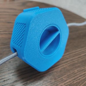 May include: A bright blue cable organizer with a hexagonal shape and a circular opening. The organizer has a textured surface and a gray cable is inserted through it. The design includes a small vent-like feature.