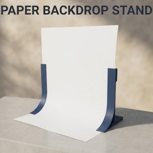 May include: A dark blue paper backdrop stand holding a single sheet of white paper. The stand features two curved side supports that hold the paper in an upright, vertical position. The text PAPER BACKDROP STAND is visible at the top of the image.