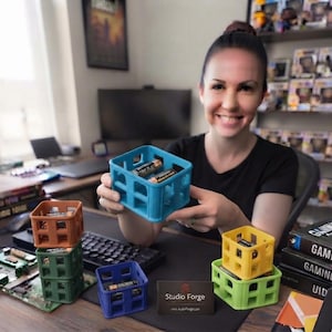 May include: A set of small, square, open-frame storage crates in blue, orange, green, yellow, and purple. Each crate features a grid-like structure with hollow sides and is designed to hold batteries. A business card reads Studio Forge.